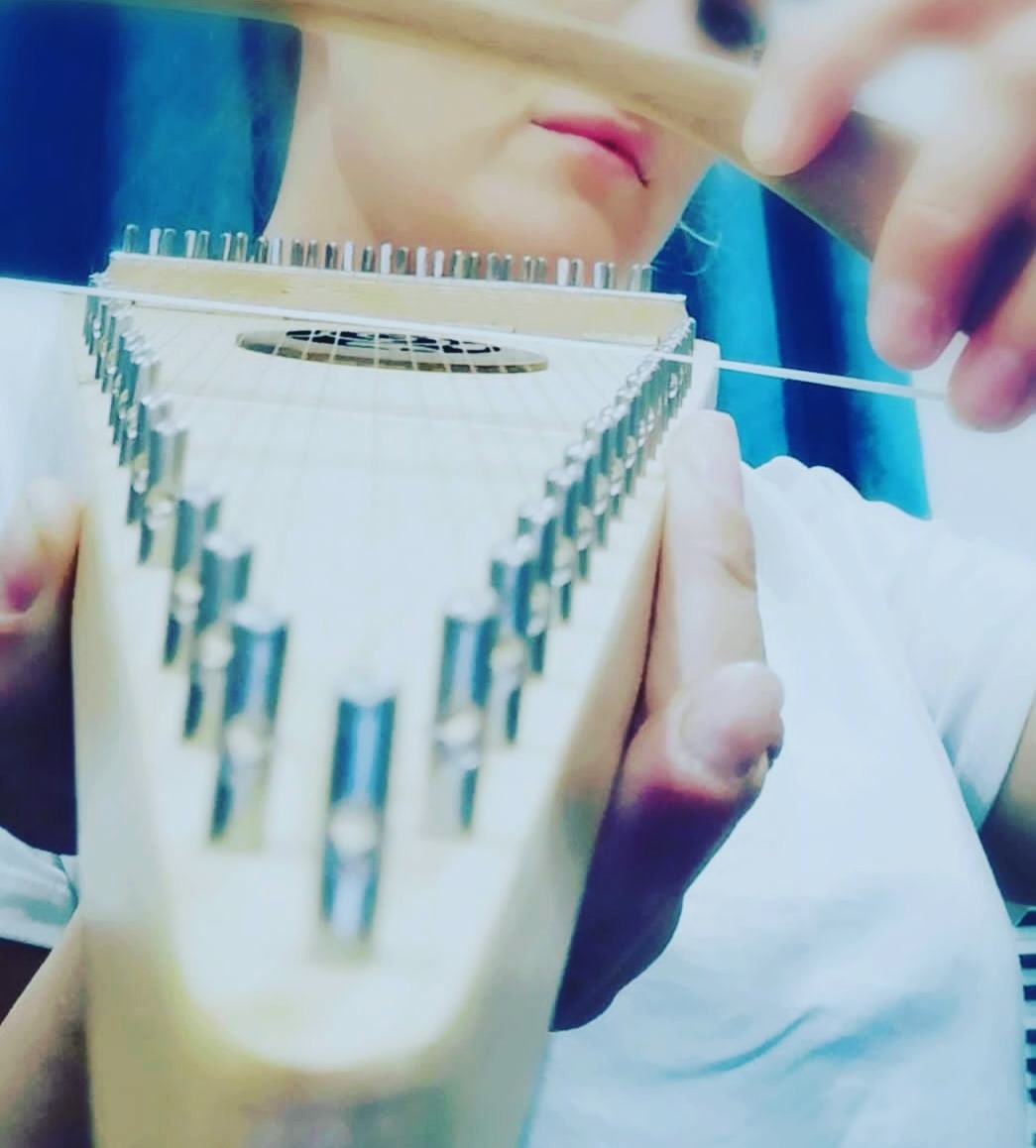 Osoba stroi autoharfę za pomocą klucza, widoczne metalowe kołki i struna naciągana na trójkątnym korpusie instrumentu.