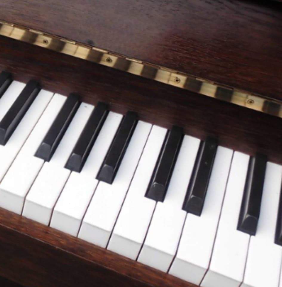 Klawiatura pianina w zbliżeniu, widoczne czarne i białe klawisze oraz fragment drewnianej obudowy z mosiężnymi elementami.