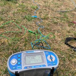 Elektrofix-Instal Cezary Pawlik - Miernik rezystancji uziemienia EurotestXE MI 3102 BT na trawie z rozciągniętymi przewodami pomiarowymi w kolorach zielonym i niebieskim, w tle widoczny wykop i torba narzędziowa.