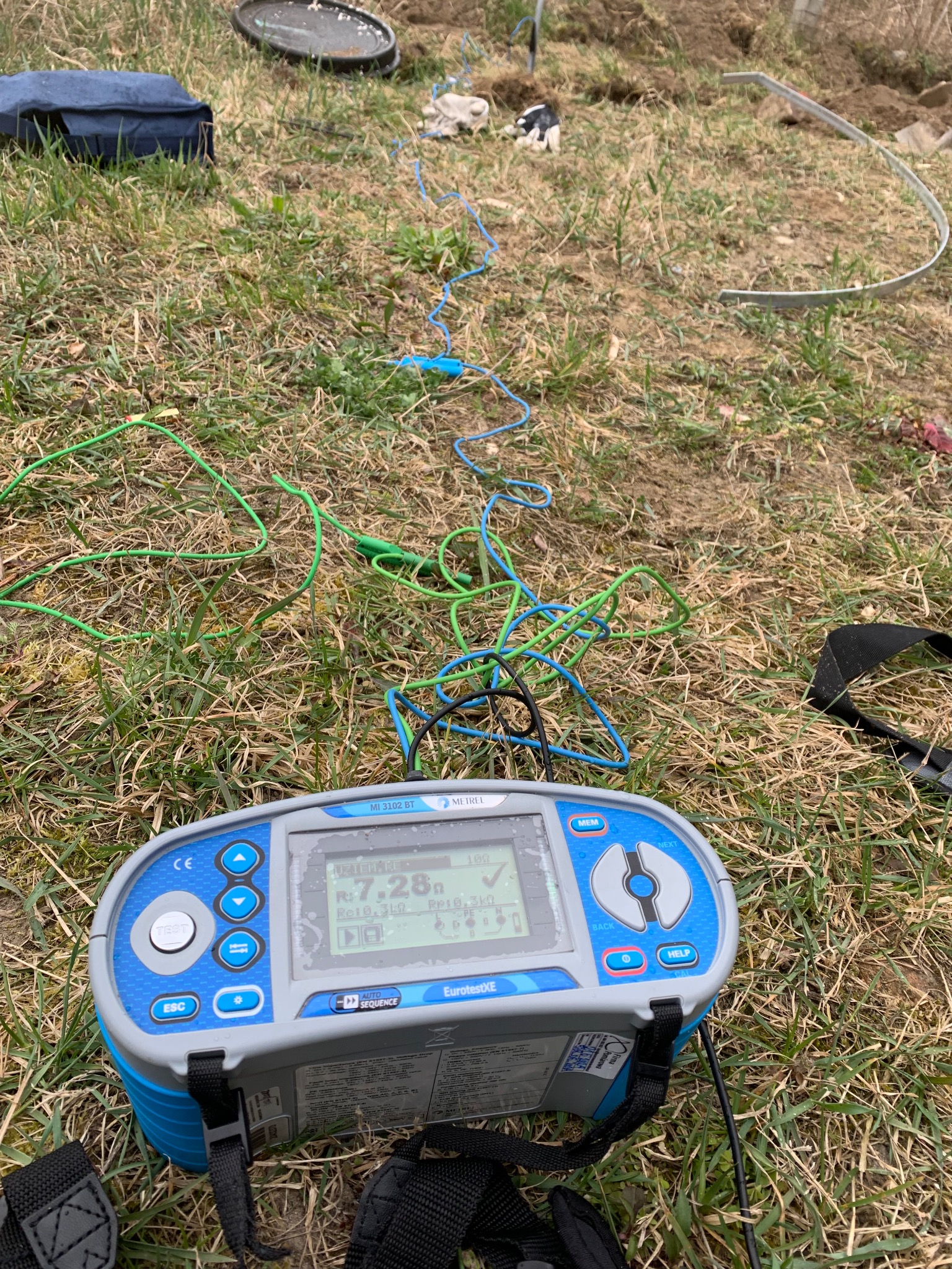 Miernik rezystancji uziemienia EurotestXE MI 3102 BT na trawie z rozciągniętymi przewodami pomiarowymi w kolorach zielonym i niebieskim, w tle widoczny wykop i torba narzędziowa.