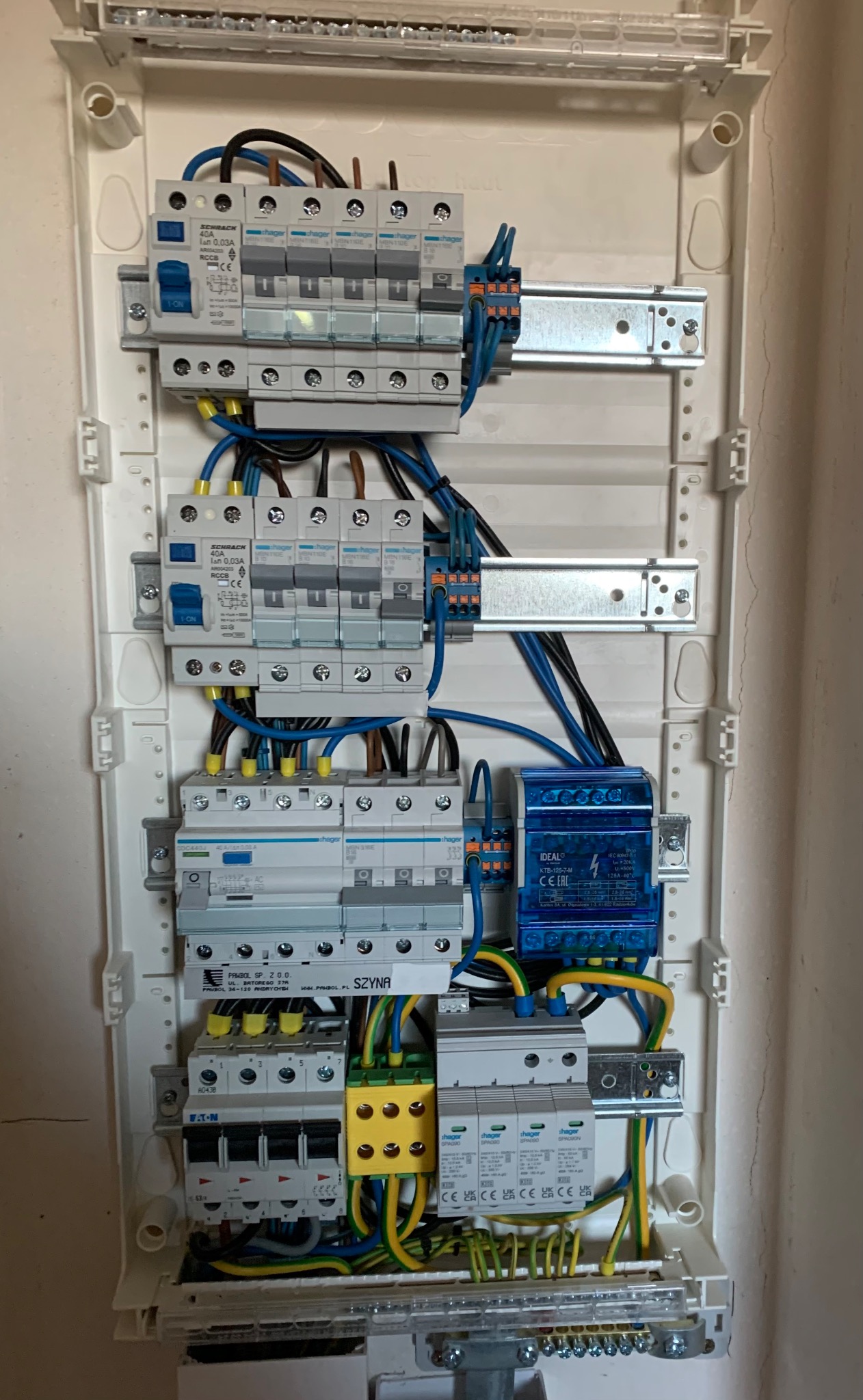 Rozdzielnica elektryczna w białej obudowie z widocznymi bezpiecznikami, wyłącznikami różnicowoprądowymi, modułem IDEAL w niebieskiej obudowie oraz okablowaniem w kolorach niebieskim, żółto-zielonym...