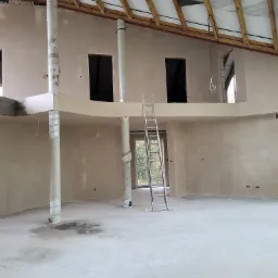 Wnętrze domu w trakcie prac wykończeniowych, widoczne świeżo nałożone tynki na ścianach, kolumny owinięte folią ochronną, metalowa drabina oparta o ścianę oraz surowe drewniane elementy konstrukcji...