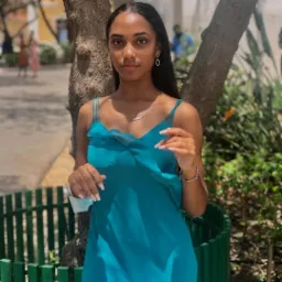 Młoda kobieta w turkusowej sukience pozuje oparta o drzewo w słoneczny dzień, z widoczną maseczką i biżuterią.