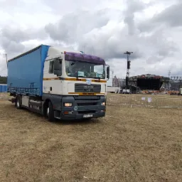 Ciężarówka MAN z niebieską plandeką zaparkowana na trawiastym terenie, w tle scena koncertowa z nagłośnieniem i oświetleniem pod pochmurnym niebem. Tablica rejestracyjna ZGR 19A1.