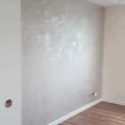 Ściana z dekoracyjną, srebrzystą fakturą w wykończonym pokoju z drewnopodobną podłogą i białymi listwami przypodłogowymi.