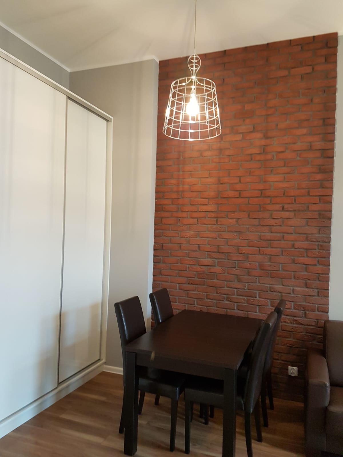 Jadalnia z ciemnym drewnianym stołem, krzesłami z brązowej skóry ekologicznej, białą szafą przesuwną i dekoracyjną ścianą z czerwonej cegły, oświetlona ażurową lampą wiszącą.