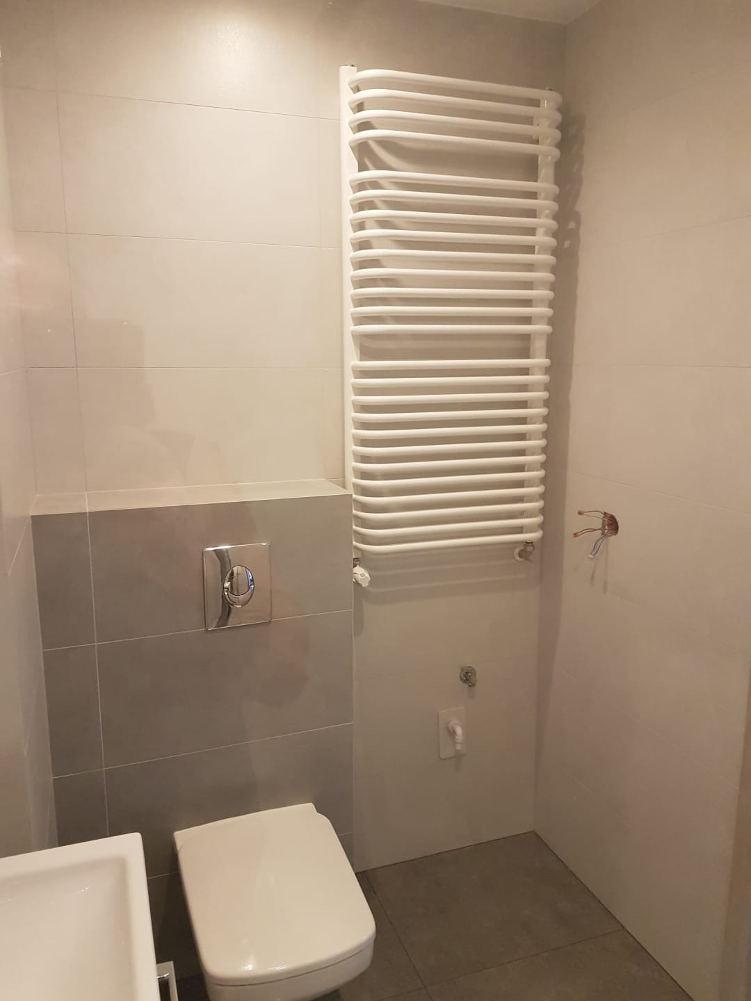 Wykończona łazienka w odcieniach szarości i bieli z grzejnikiem drabinkowym, podwieszaną toaletą i umywalką, widoczne niedokończone podłączenie elektryczne.