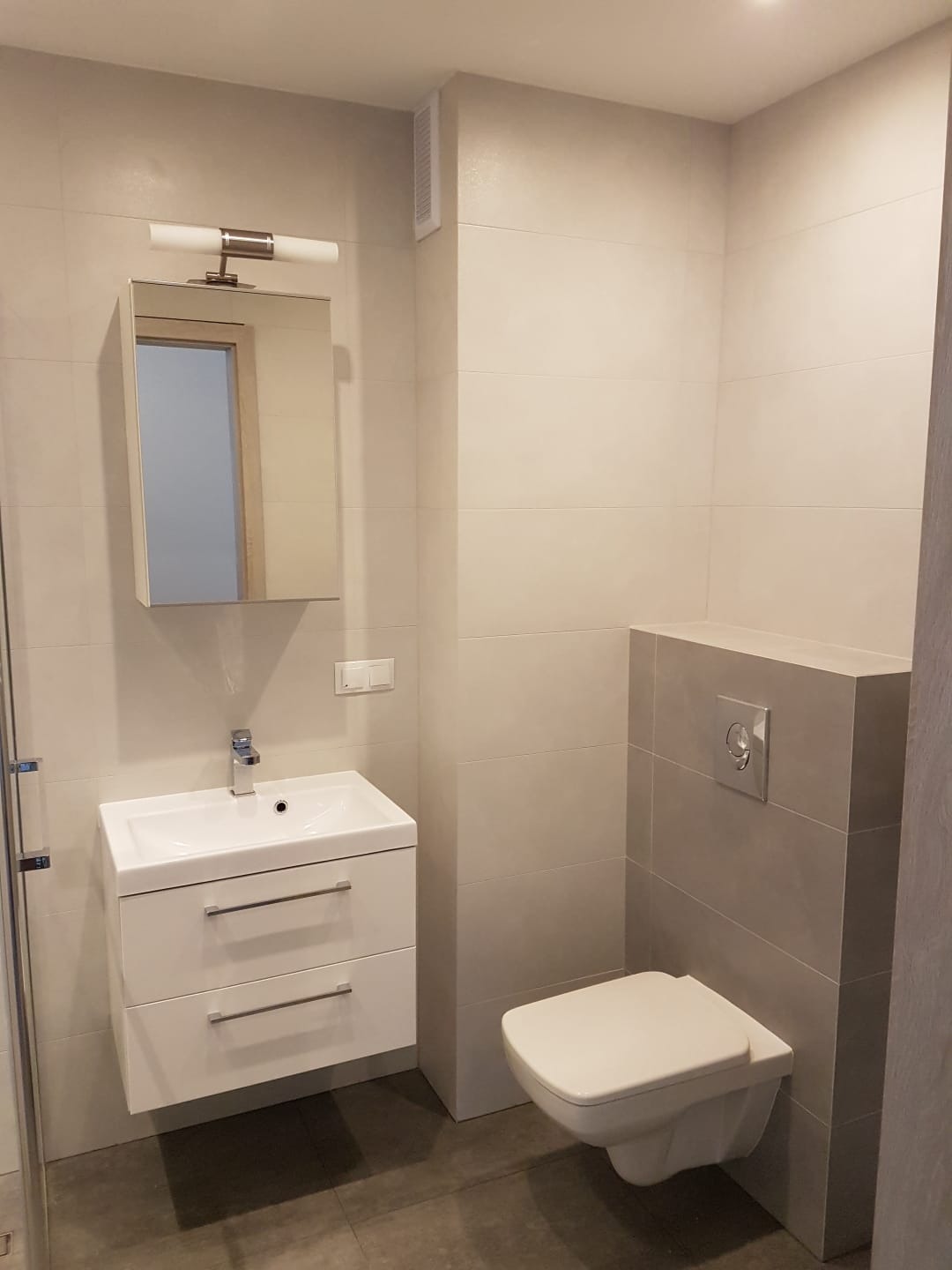 Nowoczesna łazienka w odcieniach szarości i bieli z wiszącą toaletą, szafką z umywalką i lustrem z oświetleniem, wykończona płytkami wielkoformatowymi z delikatnym połyskiem.