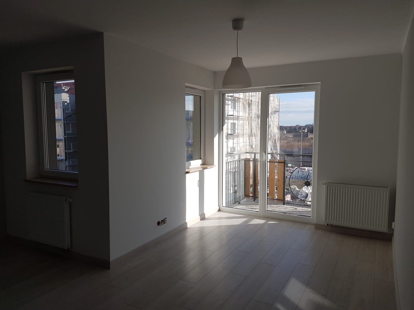 Pusty, jasny pokój z parkietem, białymi ścianami, grzejnikami i oknem balkonowym z widokiem na rusztowania budynku.