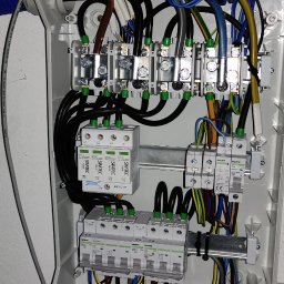 ELDANO DAWID NOWACKI - Wnętrze skrzynki elektrycznej z zainstalowanymi bezpiecznikami, ogranicznikami przepięć i przewodami w różnych kolorach, zamocowanymi na szynie DIN, widoczne oznaczenia producentów Simtec i Noark.