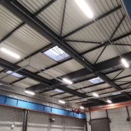 Wnętrze hali produkcyjnej z widoczną konstrukcją stropu z belek stalowych, oświetleniem jarzeniowym i świetlikami dachowymi. Widoczna suwnica i fragment ściany z blachy trapezowej.