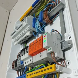 Rozdzielnica elektryczna w białej obudowie, widoczne zabezpieczenia przepięciowe w kolorze pomarańczowym, listwy zaciskowe w różnych kolorach oraz kable ułożone wewnątrz, zamontowana na ścianie...