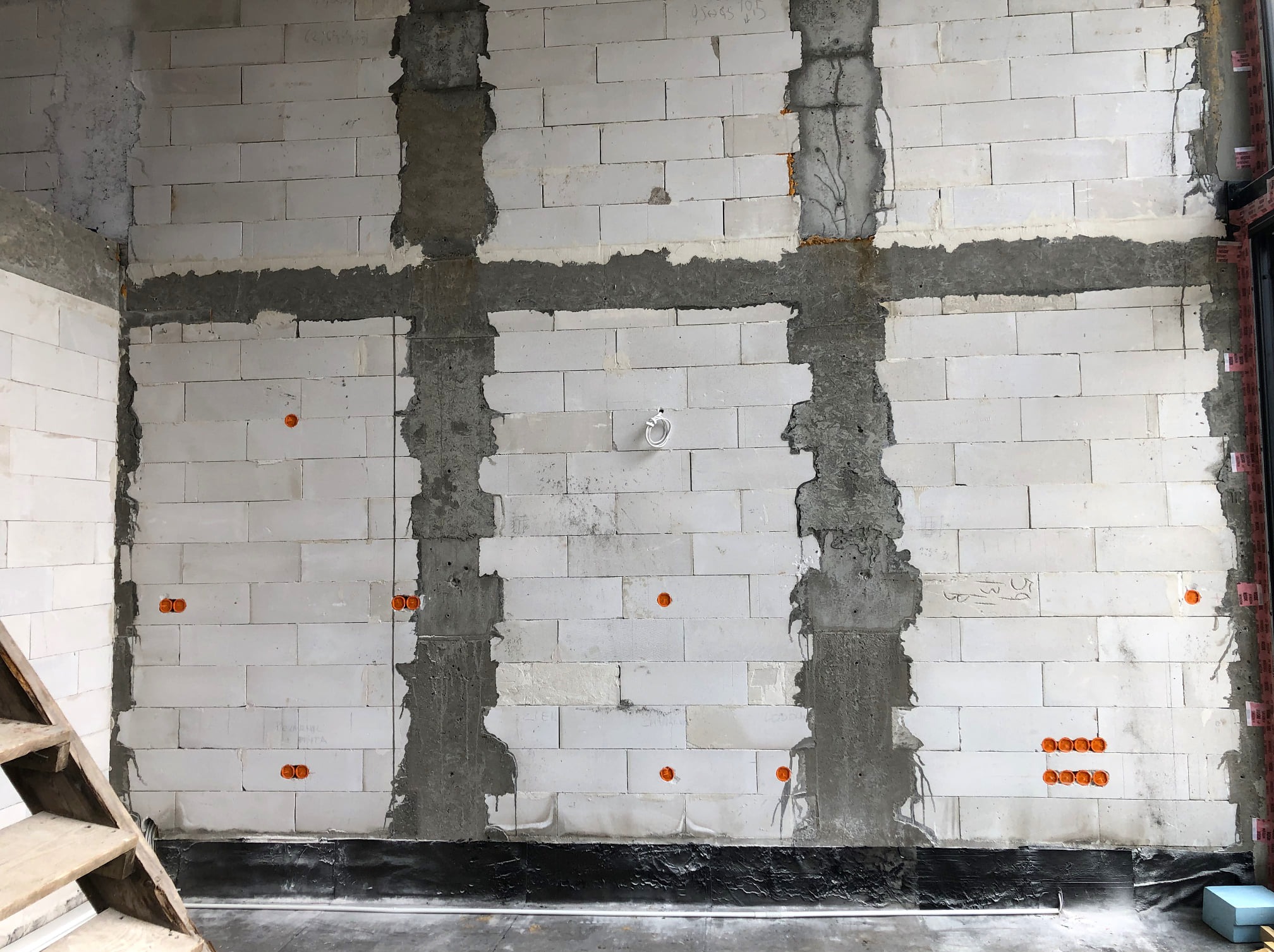 Ściana z betonu komórkowego w trakcie prac instalacyjnych, widoczne bruzdy wypełnione zaprawą, pomarańczowe puszki elektryczne w różnych miejscach ściany, biały przewód elektryczny wystający ze...