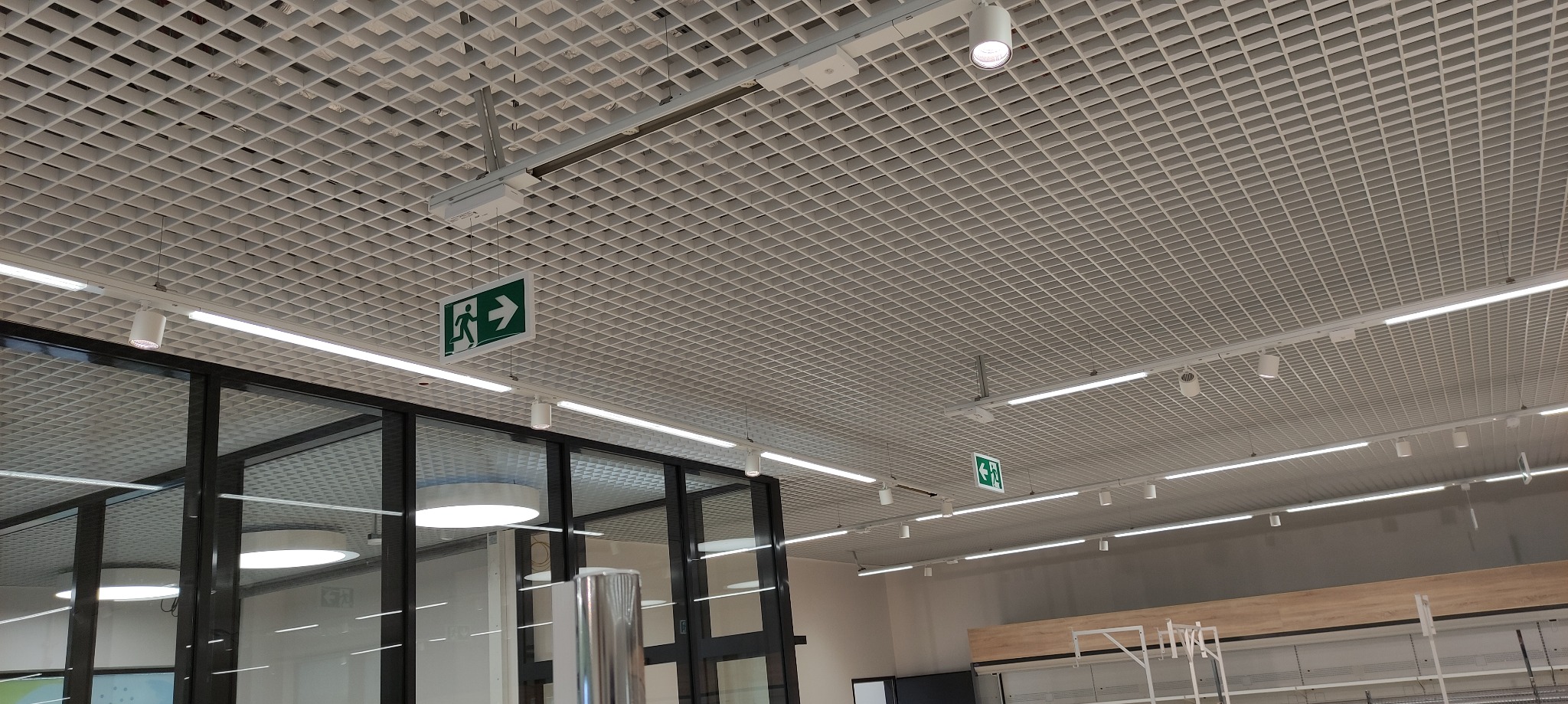 Sufit rastrowy z liniowym oświetleniem LED i punktowymi reflektorami, widoczne znaki ewakuacyjne.