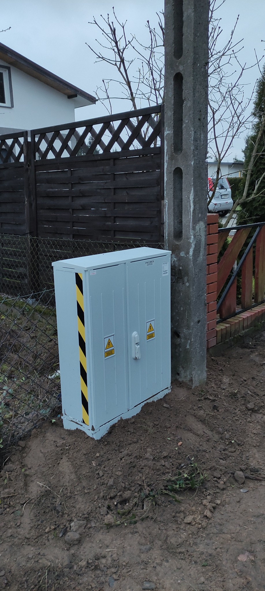 Szara skrzynka elektryczna z ostrzegawczymi znakami wysokiego napięcia, umieszczona na ziemi obok betonowego słupa energetycznego i drewnianego ogrodzenia.