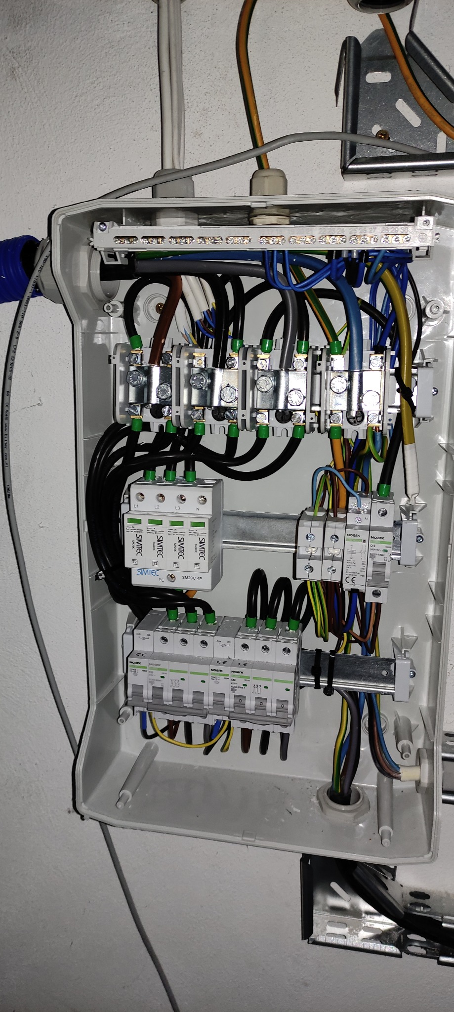 Wnętrze skrzynki elektrycznej z zainstalowanymi bezpiecznikami, ogranicznikami przepięć i przewodami w różnych kolorach, zamocowanymi na szynie DIN, widoczne oznaczenia producentów Simtec i Noark.