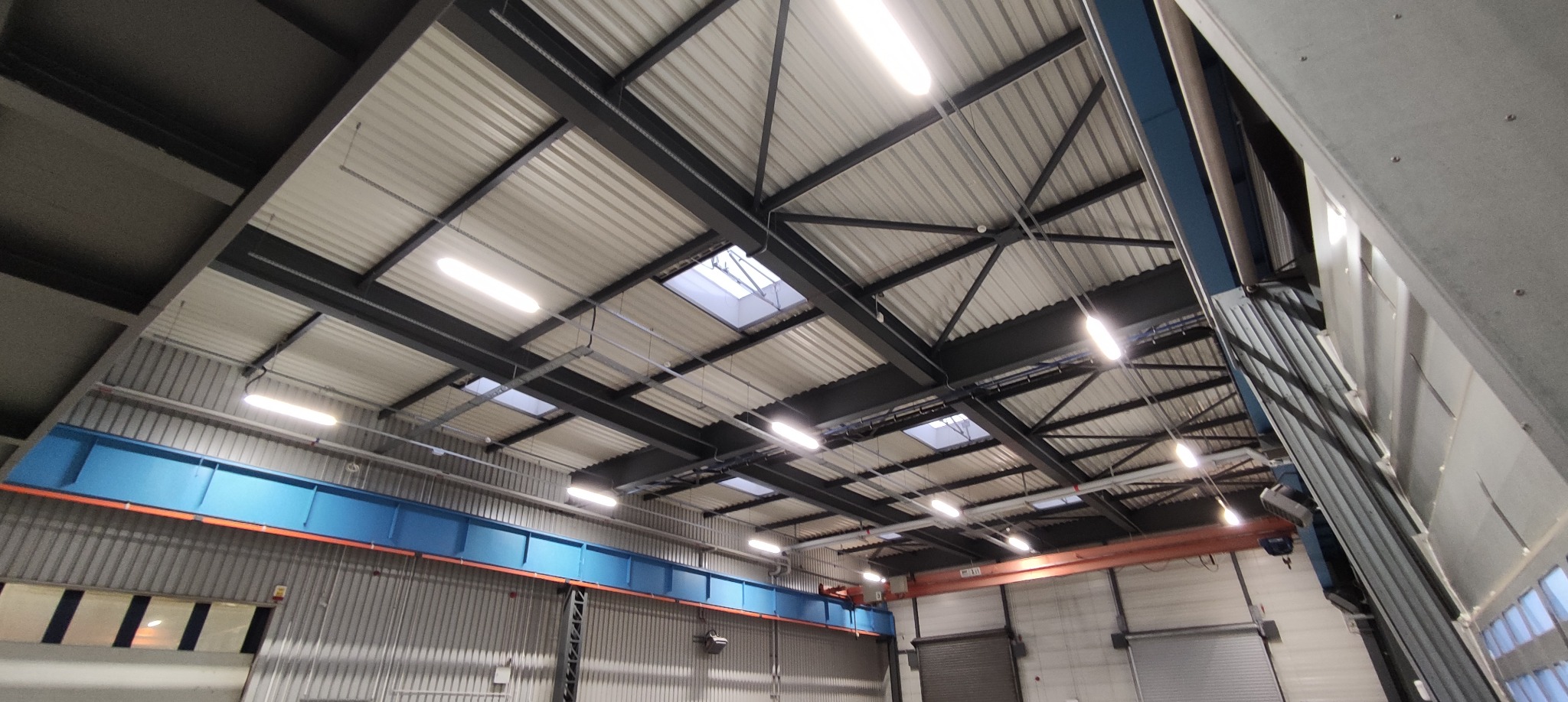 Wnętrze hali produkcyjnej z widoczną konstrukcją stropu z belek stalowych, oświetleniem jarzeniowym i świetlikami dachowymi. Widoczna suwnica i fragment ściany z blachy trapezowej.