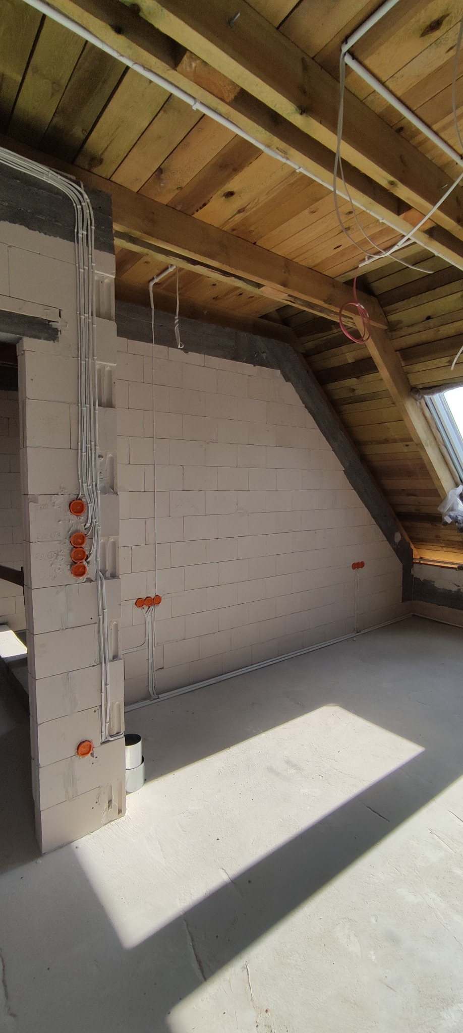 Surowy stan poddasza z widocznymi przewodami elektrycznymi w białych rurkach, pomarańczowymi puszkami instalacyjnymi w ścianie z białych bloczków oraz drewnianą konstrukcją dachu, oświetlonego...