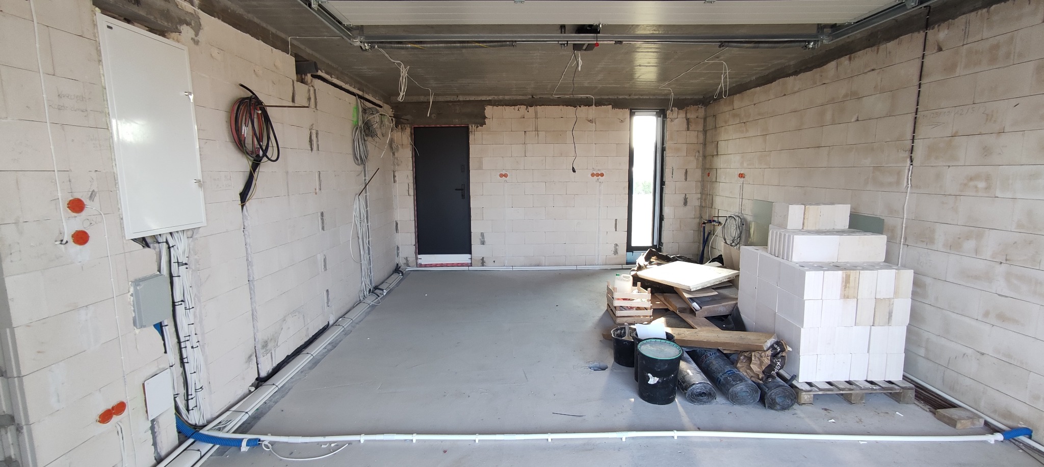 Wnętrze surowego garażu w trakcie prac instalacyjnych: widoczna biała skrzynka rozdzielcza z licznymi kablami, rury instalacyjne prowadzone wzdłuż ścian, otwory pod gniazdka, materiały budowlane...