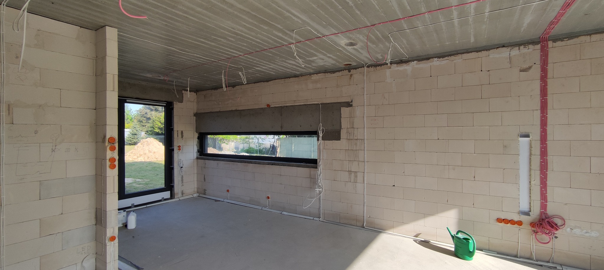 Wnętrze w stanie surowym w Kole, z widocznymi przewodami elektrycznymi prowadzonymi po ścianach i suficie, oraz puszkami instalacyjnymi. Widoczne okno i drzwi balkonowe z widokiem na zewnątrz.