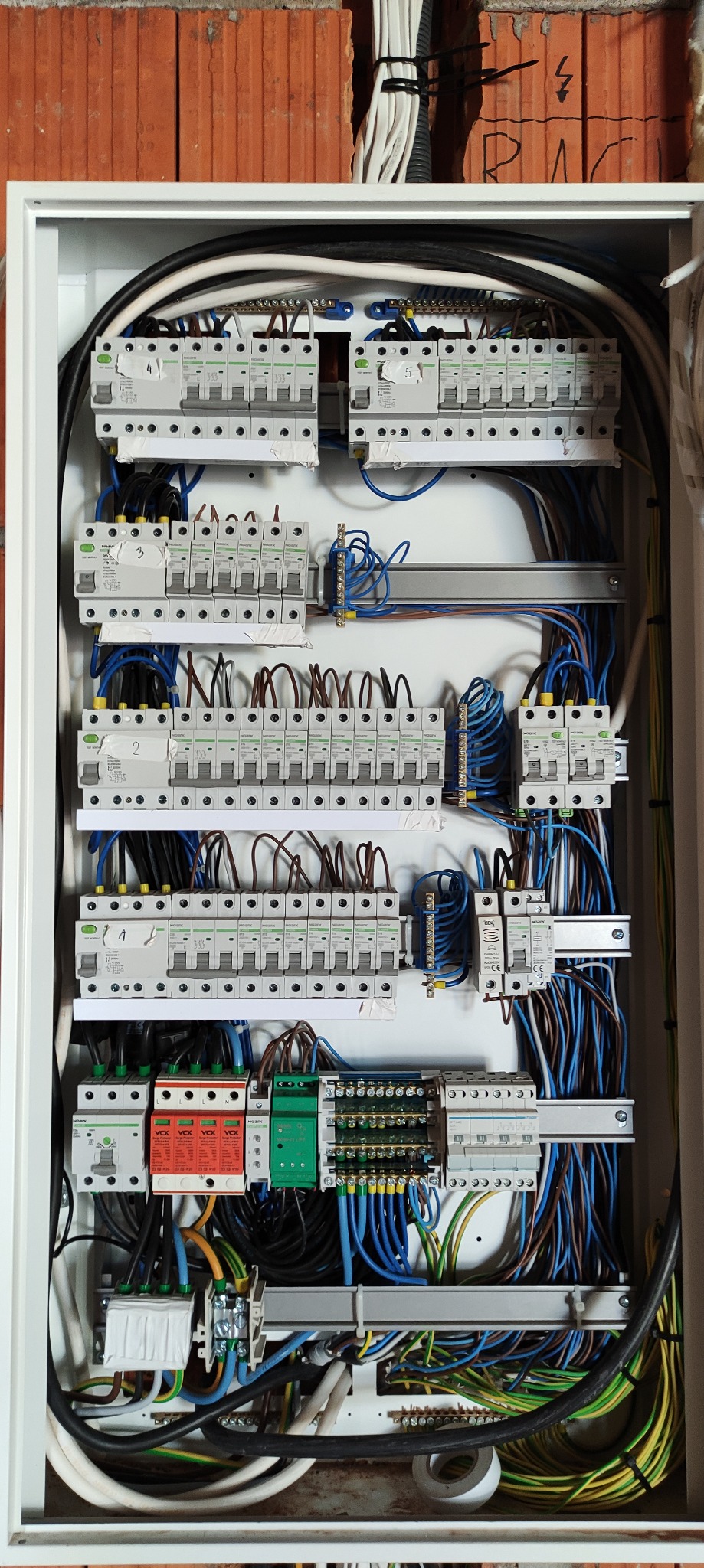 Otwarta skrzynka rozdzielcza z widocznymi zabezpieczeniami, przewodami i listwami montażowymi, zamontowana na ceglanej ścianie.