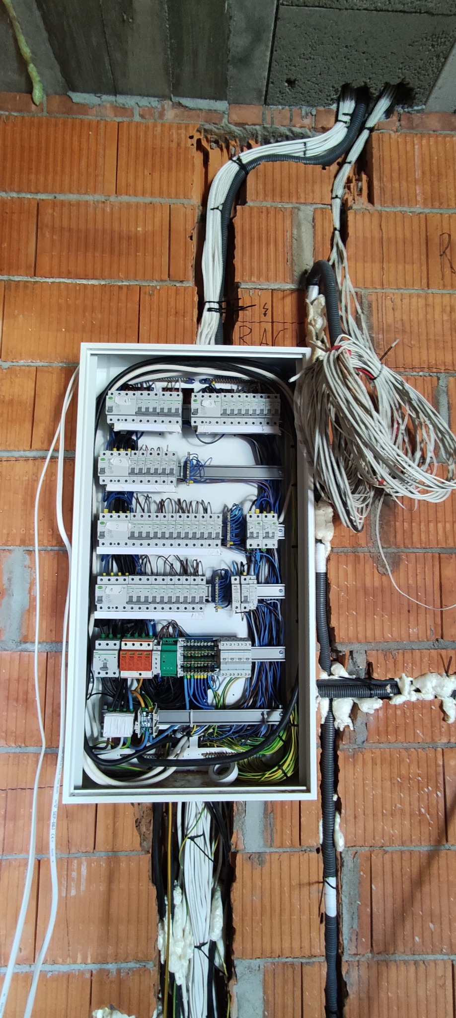Rozdzielnica elektryczna z widocznymi bezpiecznikami i przewodami, zamontowana na ścianie z czerwonej cegły w trakcie instalacji, z pianką montażową uszczelniającą przejścia kablowe.