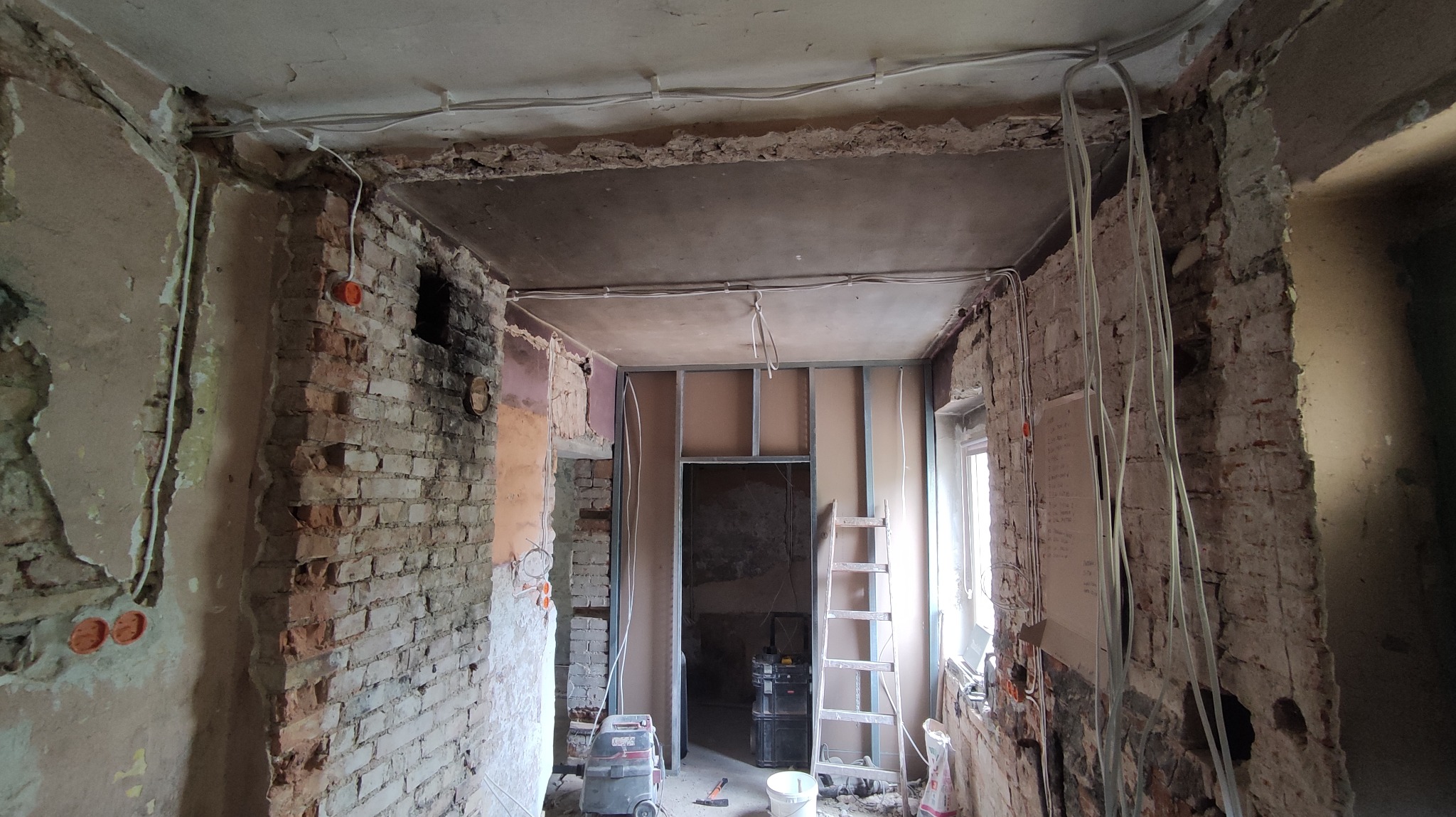 Wnętrze w trakcie remontu: odsłonięta cegła, nowe przewody elektryczne, metalowa konstrukcja ścianki działowej, drabina oparta o ścianę, widoczny sprzęt budowlany i narzędzia.