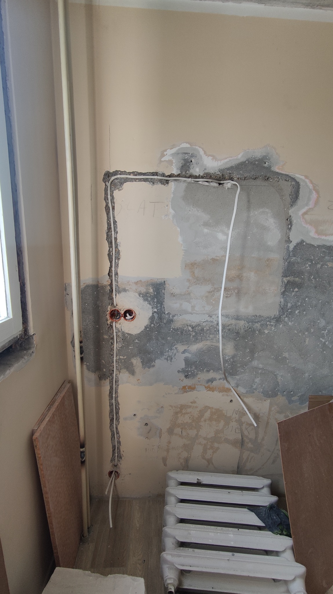 Ściana w trakcie remontu z wyżłobieniami na kable elektryczne, widoczne puszki pod gniazdka, rurka instalacyjna i fragment grzejnika.