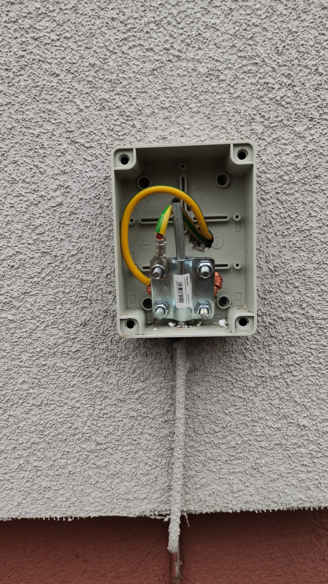 Otwarta puszka elektryczna na szorstkiej, pomalowanej ścianie, z widocznym uziemieniem, przewodem żółto-zielonym oraz metalowym zaciskiem z nakrętkami.