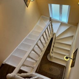 Lisa coffee Oleksander Yermolenko - Widok z góry na białe, spiralne schody z drewnianą balustradą, oświetlone lampami ściennymi i światłem naturalnym z okna dachowego, z dywanem na podeście.