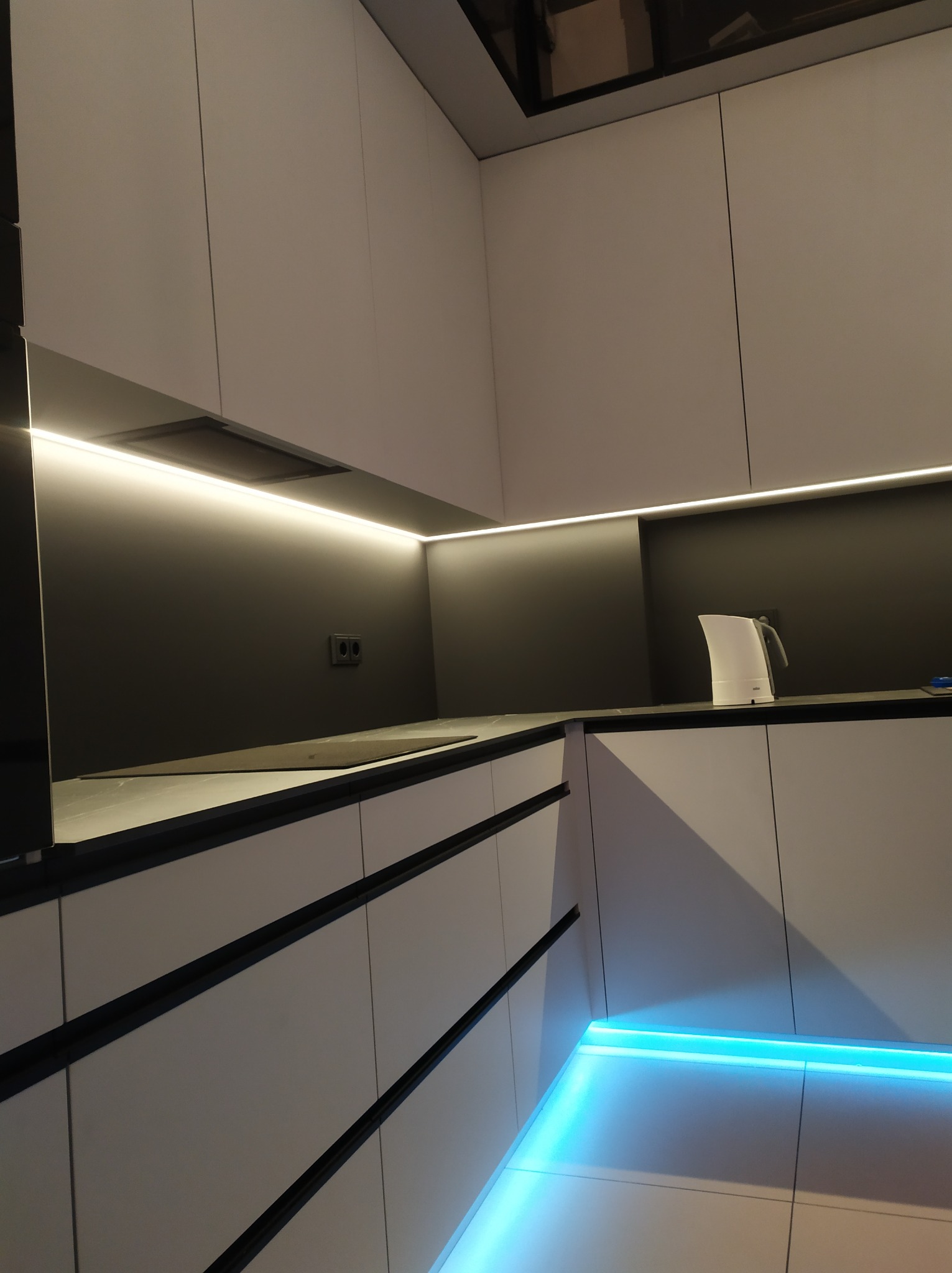 Nowoczesna kuchnia z białymi szafkami, czarnym blatem i podświetleniem LED: górne oświetlenie podszafkowe i niebieskie oświetlenie dolne, minimalistyczny czajnik na blacie.