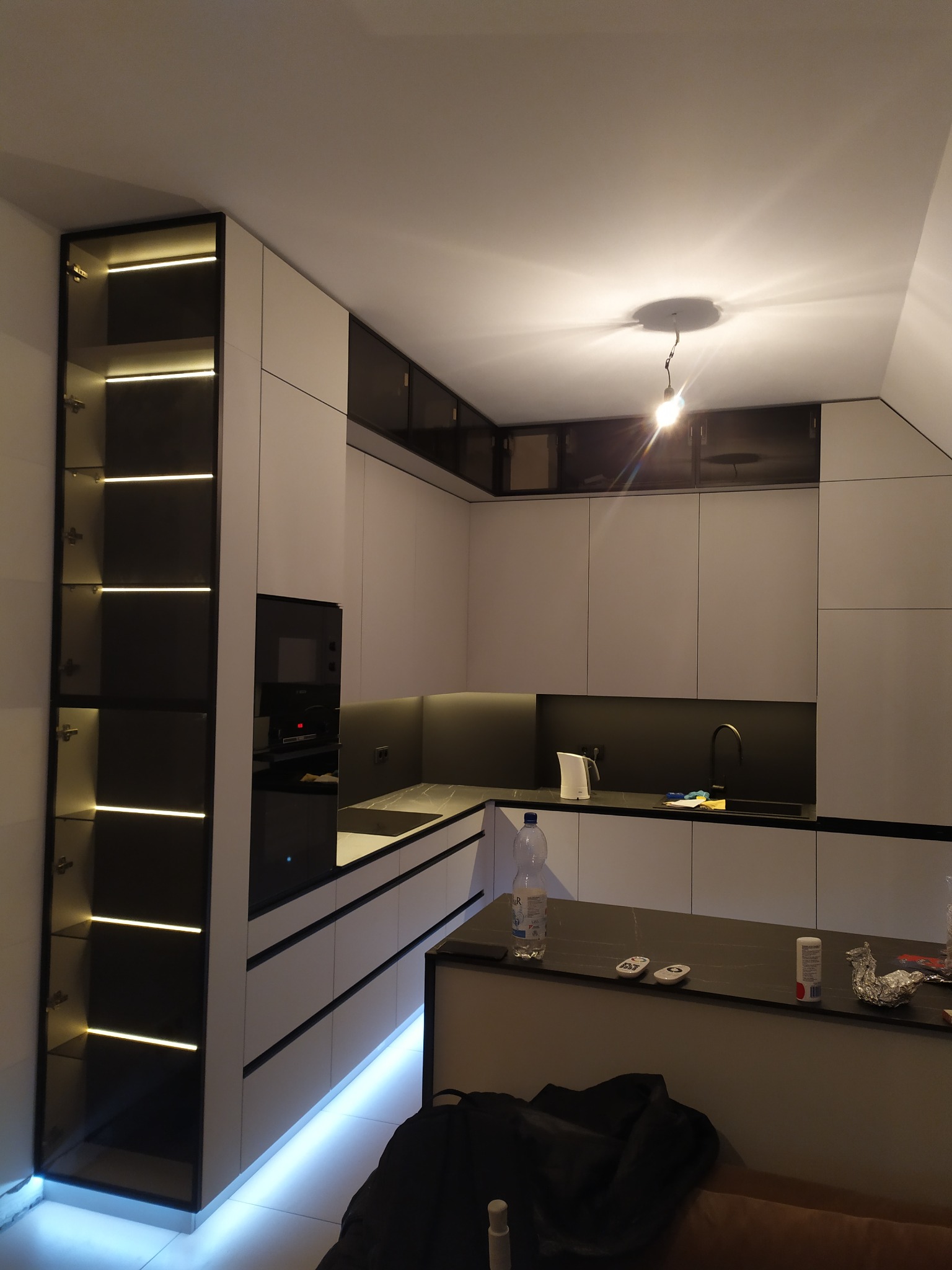 Nowoczesna kuchnia z białymi szafkami, czarnym blatem i podświetleniem LED, wysoka szafka z przeszklonymi drzwiami i oświetleniem wewnętrznym, widoczne drobne przedmioty na blacie wyspy.