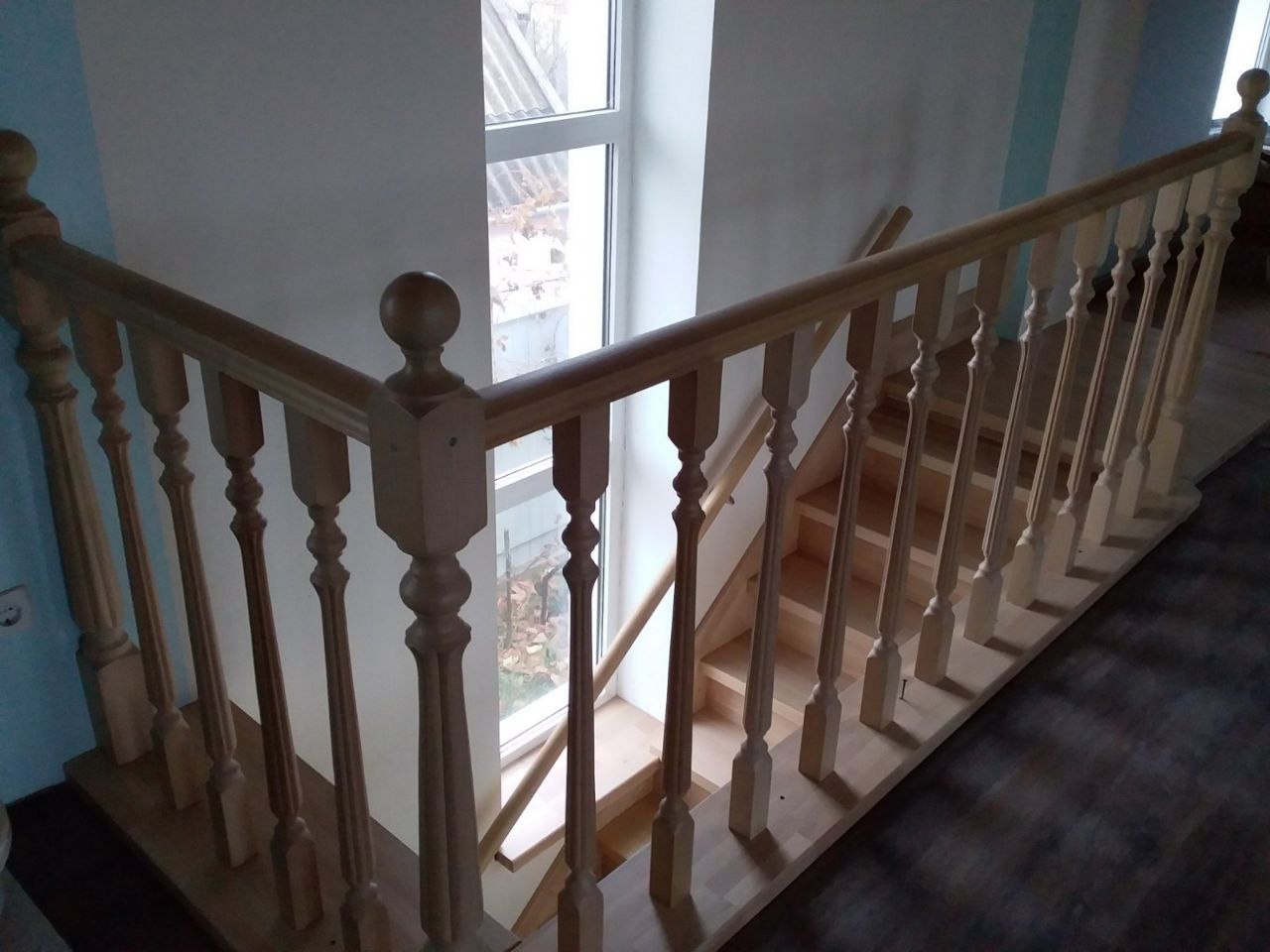 Jasne, drewniane schody z ozdobnymi balustradami, widoczne z góry, przy oknie. Podłoga częściowo widoczna, w odcieniach brązu.