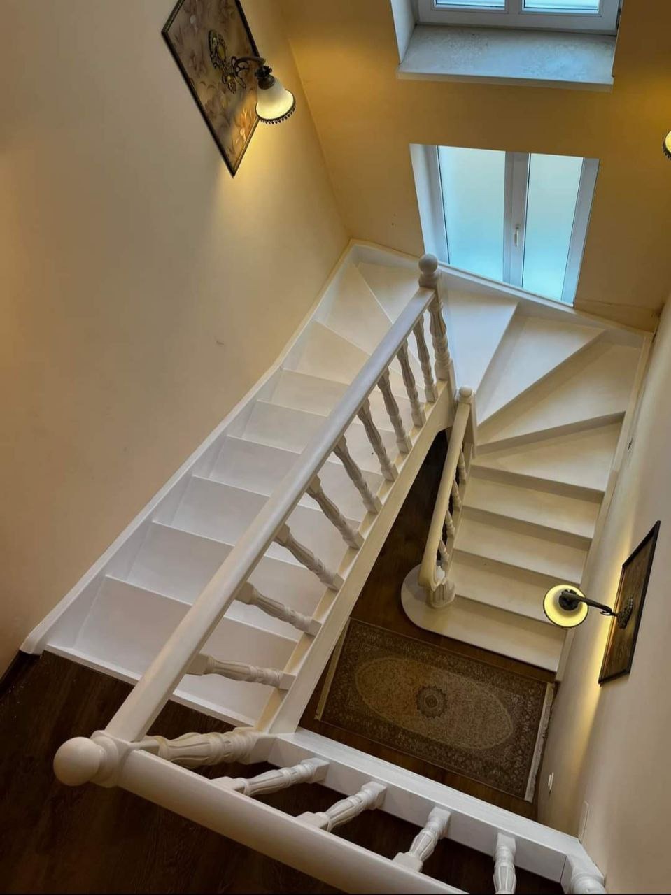 Widok z góry na białe, spiralne schody z drewnianą balustradą, oświetlone lampami ściennymi i światłem naturalnym z okna dachowego, z dywanem na podeście.