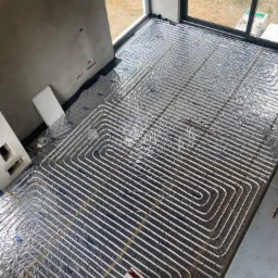 Rozłożone rury ogrzewania podłogowego na folii aluminiowej z widocznymi elementami montażowymi, w surowym wnętrzu z oknami i betonową ścianą.