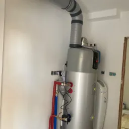 Szary, cylindryczny zasobnik ciepłej wody użytkowej z podłączonymi rurami w kolorach czerwonym i niebieskim, oraz izolowanym kominem odprowadzającym spaliny, zamontowany w surowym pomieszczeniu...