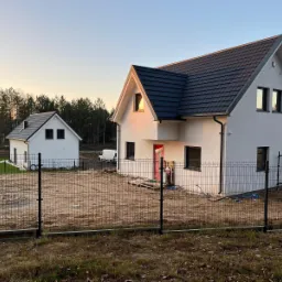 Nowoczesny, biały dom z ciemną dachówką, widoczny zza metalowego ogrodzenia, z mniejszym budynkiem gospodarczym w tle i lasem na horyzoncie, sfotografowany o zachodzie słońca.