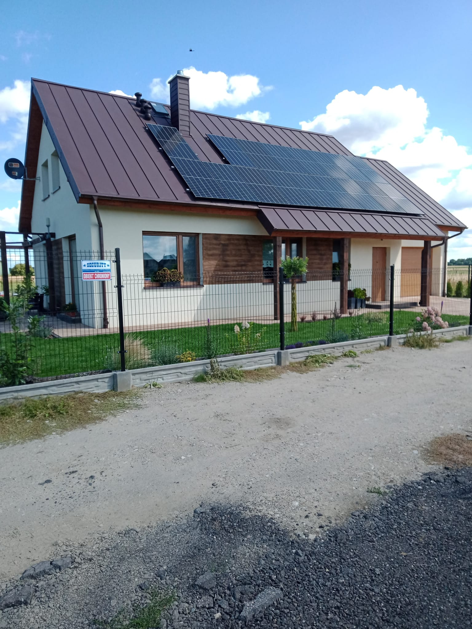 Gotowy dom jednorodzinny z brązowym dachem, panelami solarnymi, jasną elewacją z drewnianymi akcentami i ogrodzeniem z siatki, widok z drogi gruntowej.