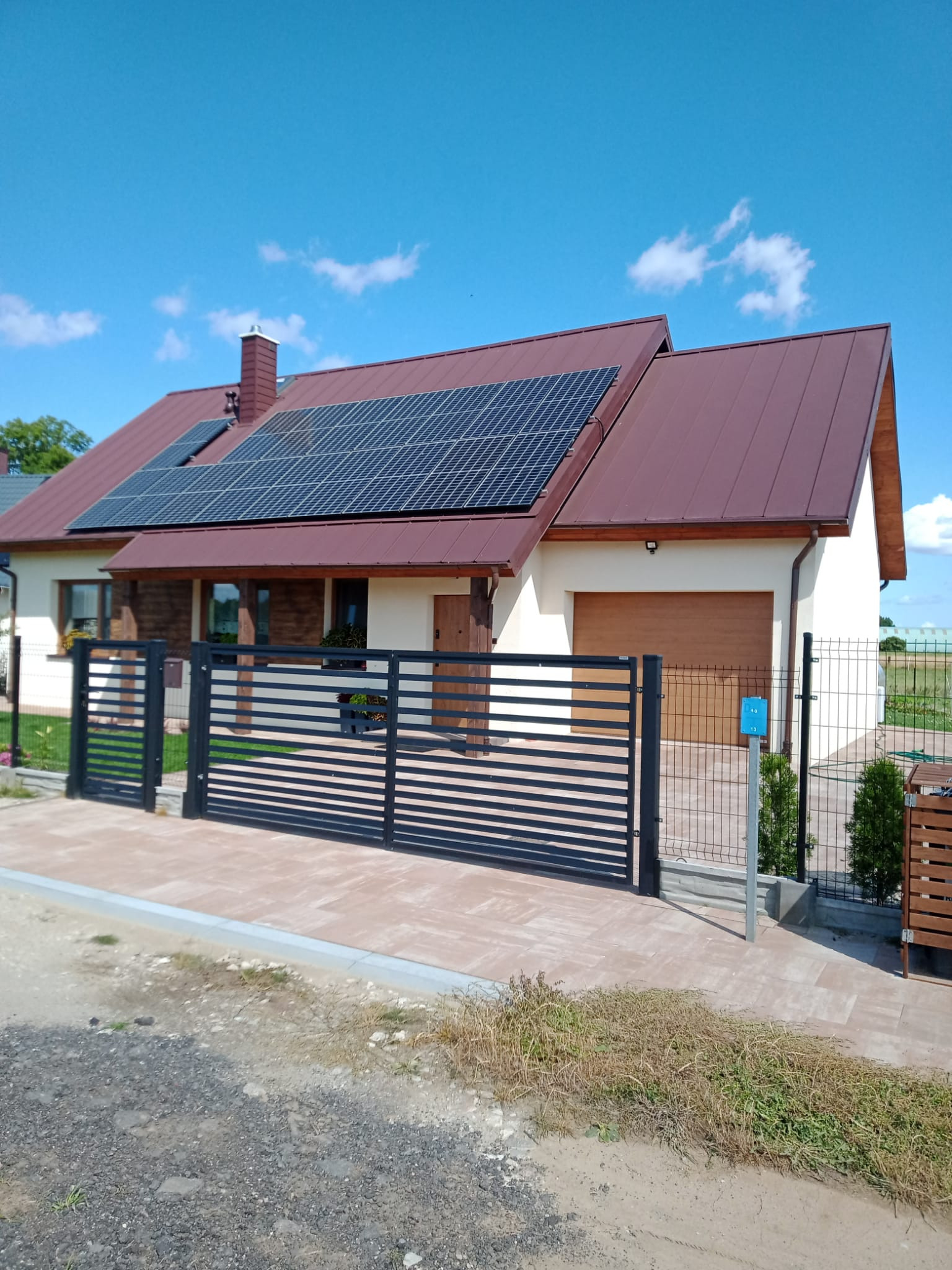 Nowoczesny dom jednorodzinny z panelami słonecznymi na dachu z brązowej blachy, ogrodzony czarnym, poziomym ogrodzeniem panelowym, z widocznym fragmentem elewacji w kolorze kremowym i elementami...