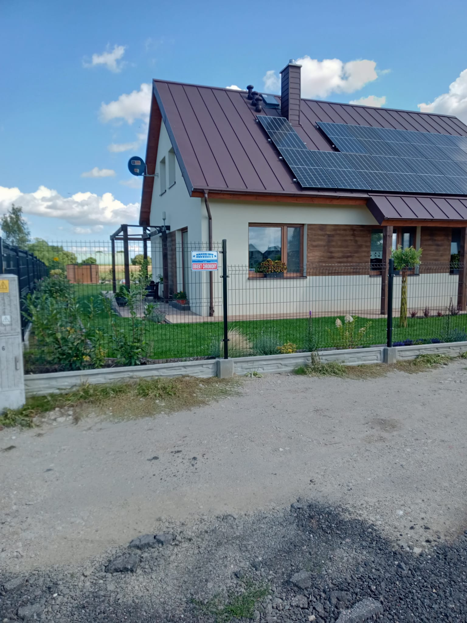 Nowoczesny dom jednorodzinny z brązowym dachem, panelami słonecznymi i elementami drewnianej elewacji, otoczony metalowym ogrodzeniem z betonową podmurówką i zadbanym trawnikiem.
