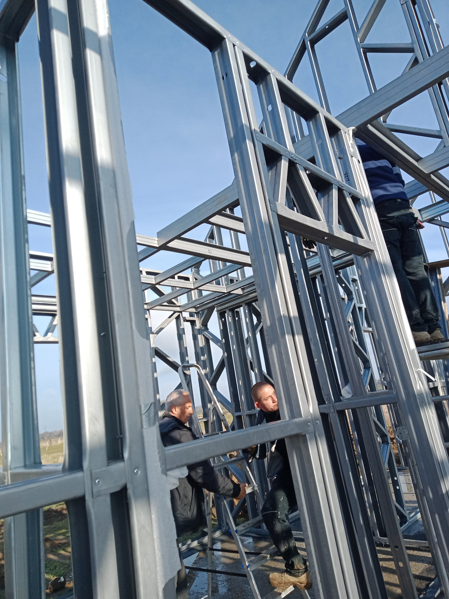 Montaż metalowej konstrukcji domu: pracownicy na drabinie mocują szkielet z profili stalowych na tle błękitnego nieba.
