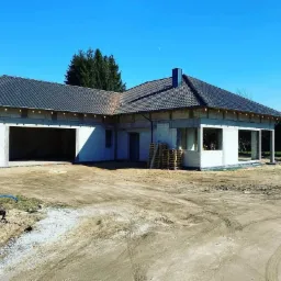 Niedokończony parterowy budynek z garażem i ciemną dachówką w słoneczny dzień, widoczne palety i kontener na gruz.