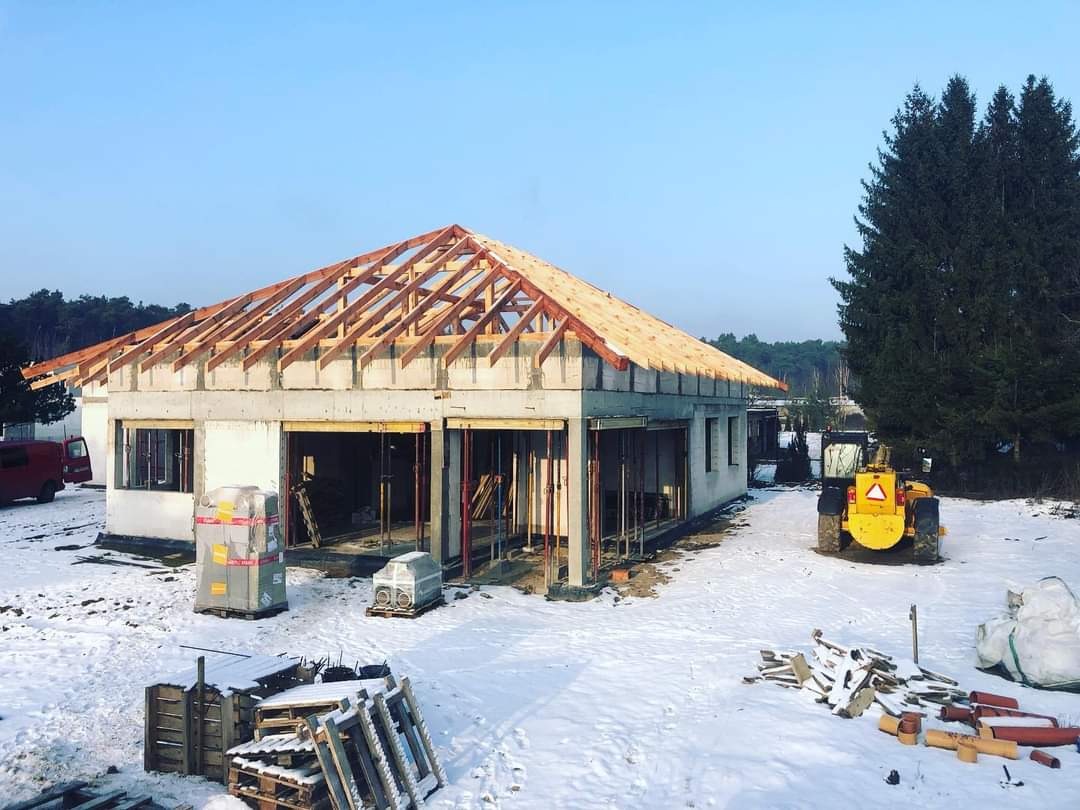 Budowa domu jednorodzinnego w zimowej scenerii: surowy stan z drewnianą konstrukcją dachu, widoczne bloczki betonowe, palety z materiałami budowlanymi, żółta ładowarka w tle.