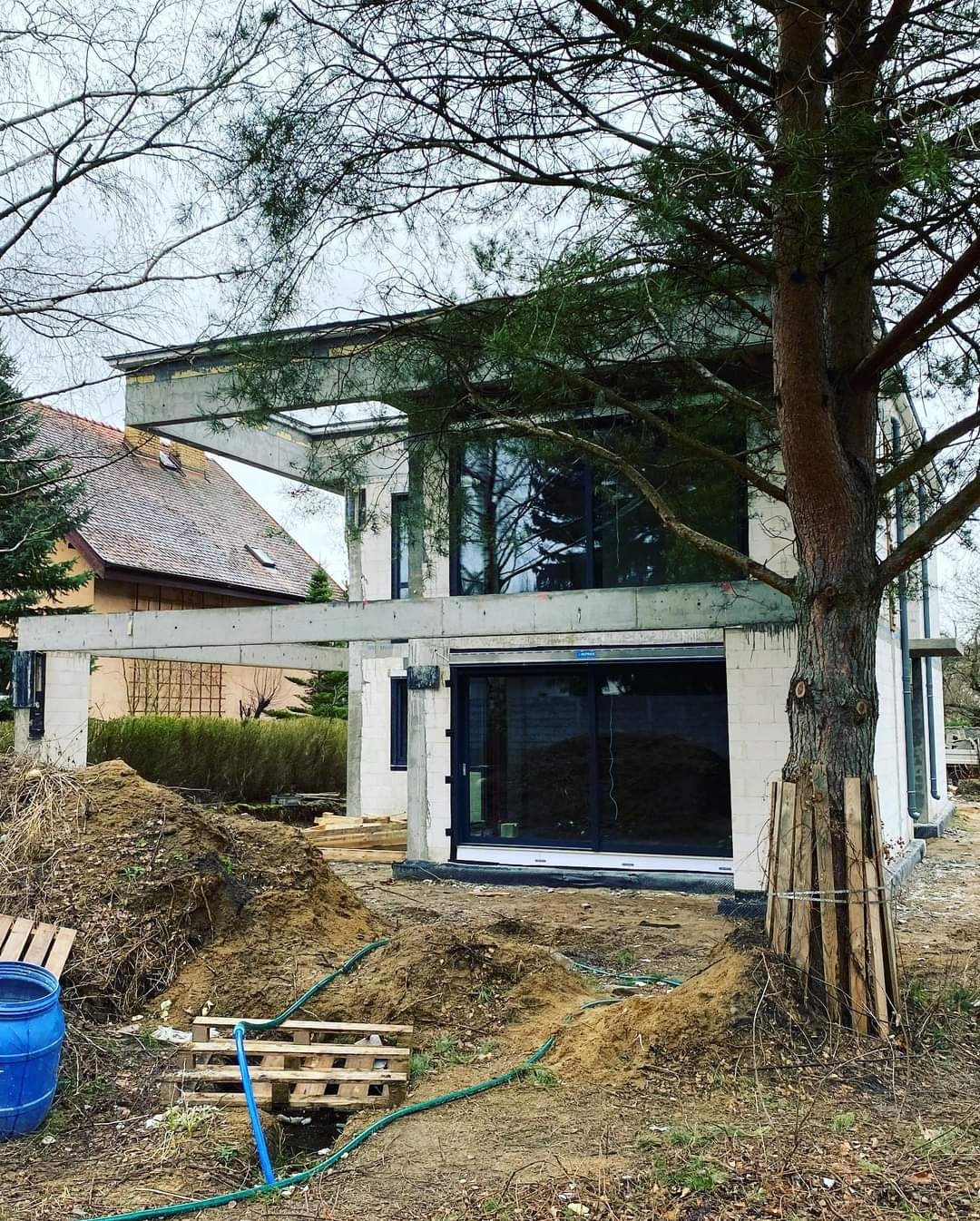 Surowy stan domu z betonu komórkowego z dużymi przesuwnymi oknami, widoczne elementy konstrukcyjne i przygotowany teren z hałdą ziemi, drzewo zabezpieczone deskami przy budowie.
