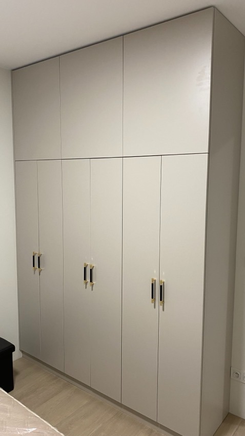 Wysoka, zabudowana szafa z jasnoszarymi frontami, wyposażona w podłużne, czarno-złote uchwyty, sięgająca od podłogi do sufitu, z widocznymi gniazdkami elektrycznymi na ścianie i fragmentem podłogi...