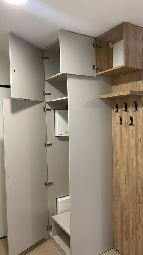 Wnętrze szafy wnękowej z otwartymi drzwiami, widoczna skrzynka elektryczna, półka na buty i panel w drewnopodobnym dekorze z wieszakami.