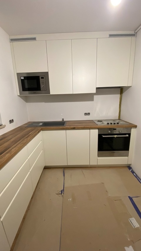Nowoczesna kuchnia z białymi frontami szafek i drewnianym blatem, w trakcie montażu, podłoga zabezpieczona kartonem, widoczne gniazdka elektryczne bez osprzętu.