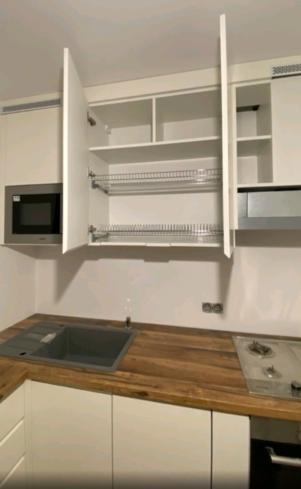 Nowoczesna kuchnia z białymi szafkami, drewnianym blatem, wbudowaną mikrofalówką i suszarką na naczynia w górnej szafce, widok z perspektywy narożnika.