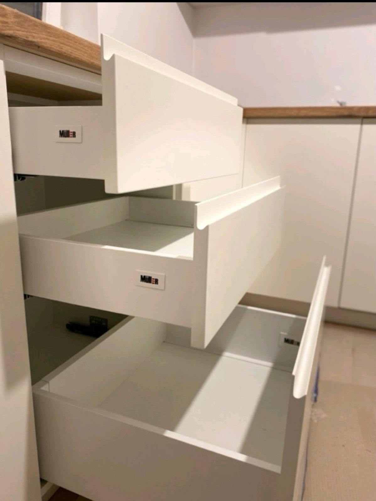 Wysuwane, białe szuflady z logo producenta w zabudowie kuchennej z drewnianym blatem, częściowo otwarte.