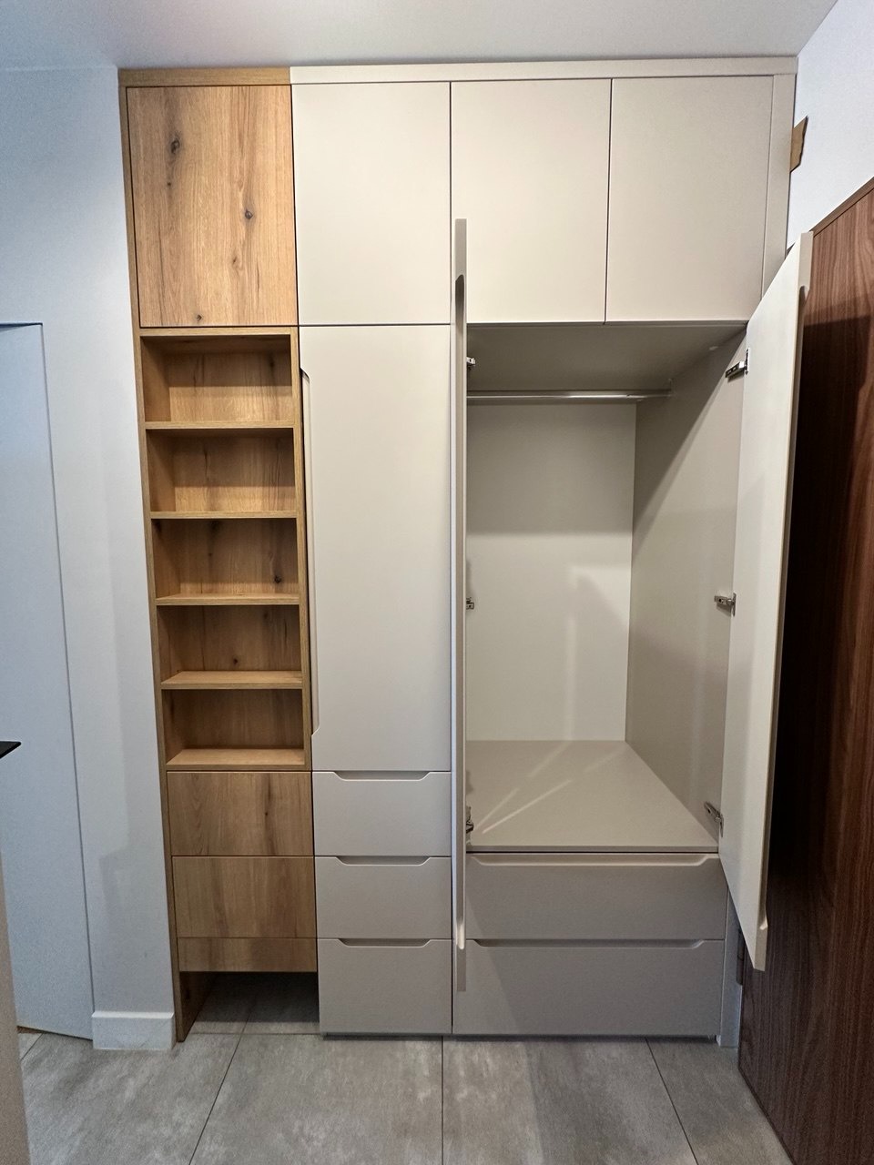 Jasna szafa wnękowa z drewnianymi akcentami, otwartymi półkami i szufladami. Jedne drzwi otwarte, ukazują wnętrze z drążkiem na ubrania. Nowoczesny design.